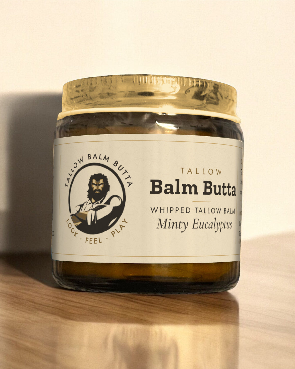 Tallow Roots Co. Minty Eucalyptus Balm Butta whipped tallow balm in a glass jar with gold lid and cream label, sitting on a wooden shelf against a plain wall