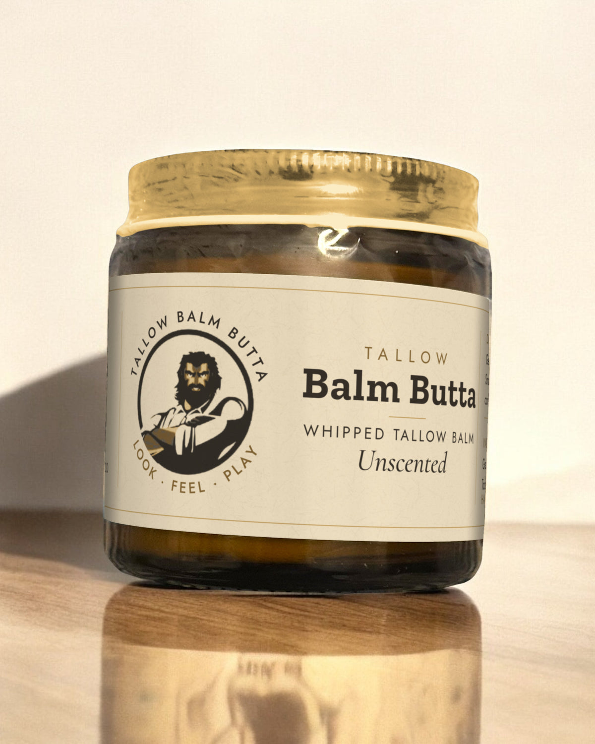 Tallow Roots Co. Unscented Balm Butta whipped tallow balm in a glass jar with gold lid and cream label, sitting on a wooden shelf against a plain wall