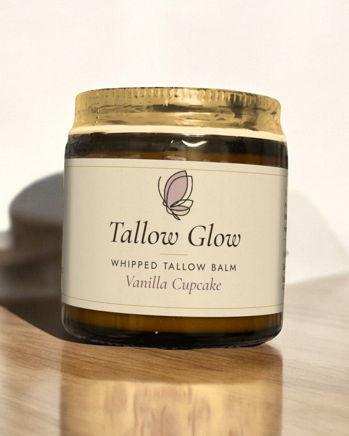 Tallow Roots Co. Vanilla Cupcake Tallow Glow whipped tallow balm in a glass jar with gold lid and cream label, sitting on a wooden shelf against a plain wall